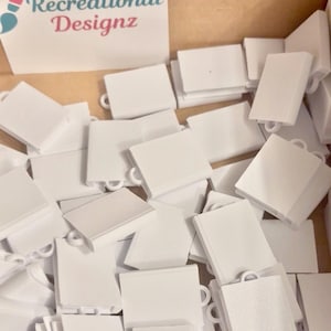 Puede incluir: Una colección de pequeños componentes rectangulares de plástico blanco, cada uno con un pequeño lazo integrado en la parte superior para colgar. Estos artículos están esparcidos en una caja junto a una tarjeta blanca con el texto Recreational Designz.