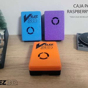 May include: Three 3D printed cases for a Raspberry Pi 3/4 computer. The cases are blue, purple, and orange. The text "VELEZ 3D" is printed on each case. The text "CAJA PARA RASPBERRY PI 3/4" is printed above the cases. The text "*SOLO CAJA, NO INCLUYE RASPBERRY" is printed below the cases.