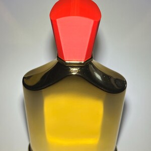 May include: A gold-colored perfume bottle with a red cap. The bottle has a rectangular shape with rounded edges and a gold base. The red cap is a geometric shape. The bottle is filled with a yellow liquid.
