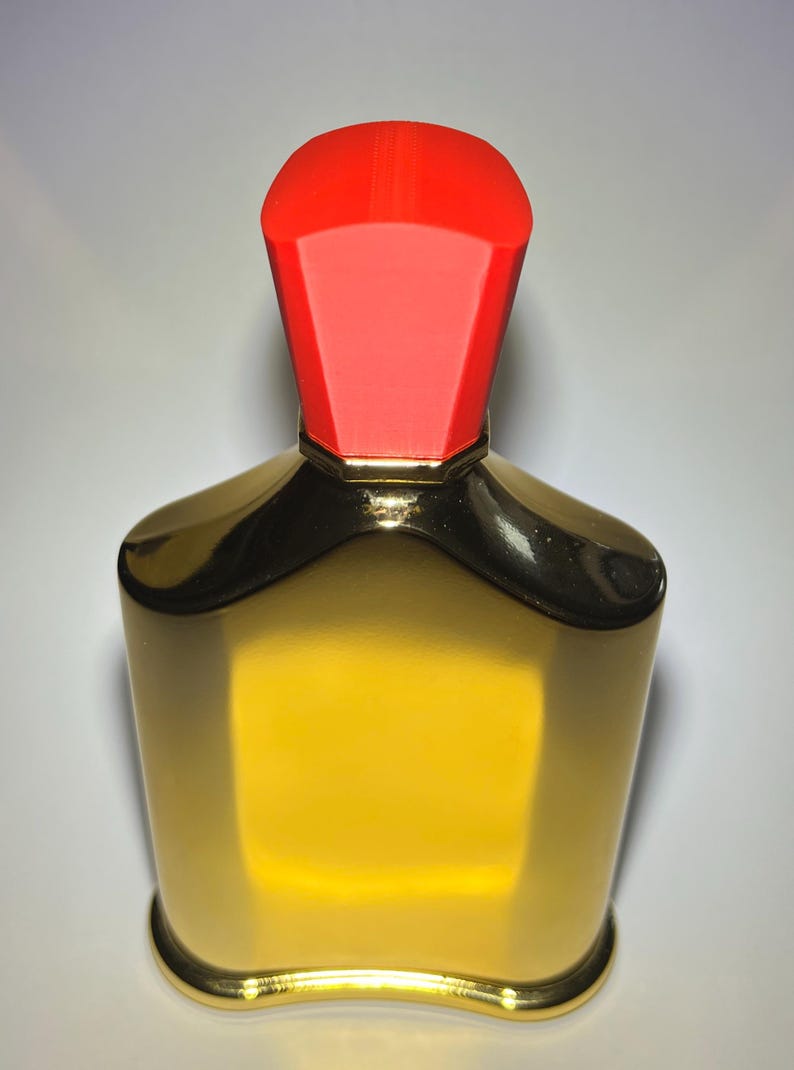 May include: A gold-colored perfume bottle with a red cap. The bottle has a rectangular shape with rounded edges and a gold base. The red cap is a geometric shape. The bottle is filled with a yellow liquid.