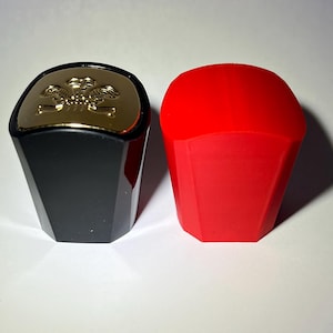 Könnte beinhalten: Zwei Flaschenverschlüsse. Einer ist schwarz mit einem goldenen Deckel mit einem dekorativen Emblem. Der andere ist einfarbig rot. Beide Verschlüsse haben ein facettiertes, geometrisches Design und sind wahrscheinlich für Wein- oder Spirituosenflaschen.