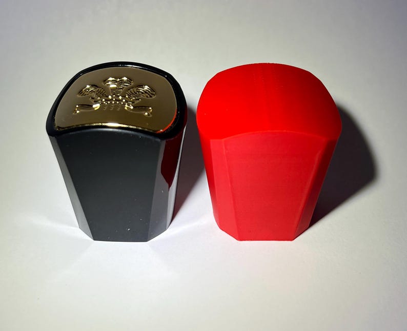 May include: Two bottle stoppers. One is black with a gold top featuring a decorative emblem. The other is a solid red color. Both stoppers have a faceted, geometric design, and are likely for wine or liquor bottles.