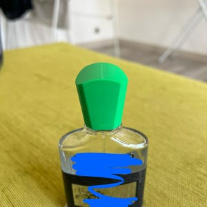 May include: A clear glass perfume bottle with a bright green cap. The bottle has a black label and is sitting on a yellow surface. The bottle is likely filled with perfume or cologne.