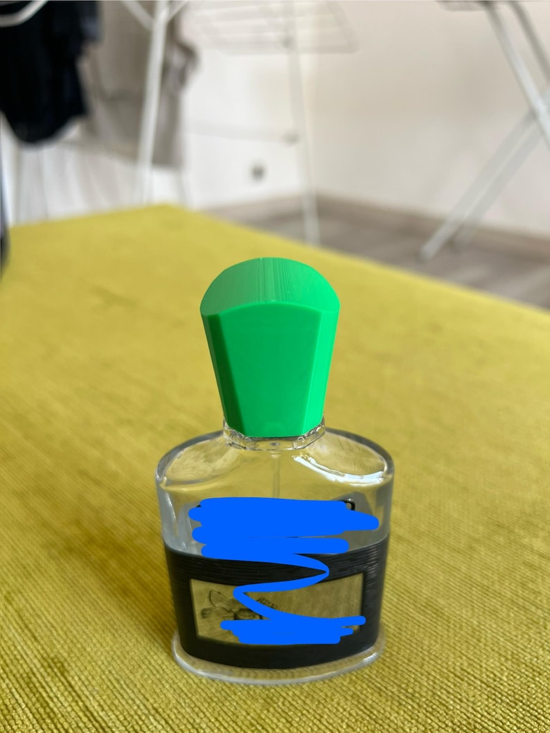 May include: A clear glass perfume bottle with a bright green cap. The bottle has a black label and is sitting on a yellow surface. The bottle is likely filled with perfume or cologne.