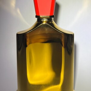 May include: A gold-colored perfume bottle with a red cap. The bottle has a rectangular shape with rounded edges and a shiny, reflective surface. The cap is a trapezoid shape.