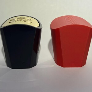May include: Two bottle stoppers. One is black with a gold top, the other is red. The stoppers have a geometric design and are likely made of plastic or a similar material. They are designed to seal bottles.