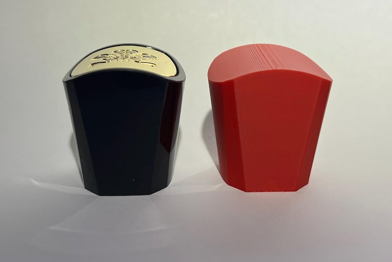 May include: Two bottle stoppers. One is black with a gold top, the other is red. The stoppers have a geometric design and are likely made of plastic or a similar material. They are designed to seal bottles.