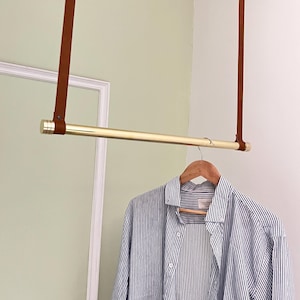 May include: A hanging clothes rail made of brass, supported by brown leather straps. A blue and white striped shirt hangs on a wooden hanger. The rail offers a space-saving storage solution.