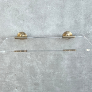 May include: A clear acrylic shelf with brass-colored metal brackets. The shelf has a rectangular shape with angled front corners. The brackets are circular and mounted to the wall. A modern and minimalist design for home decor.