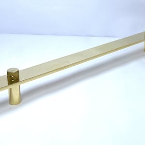 Pode incluir: Um puxador de armário dourado com uma barra retangular e dois postes cilíndricos. A ferragem é feita de metal e tem um acabamento polido. O puxador é projetado para uso em armários, gavetas ou outros móveis.