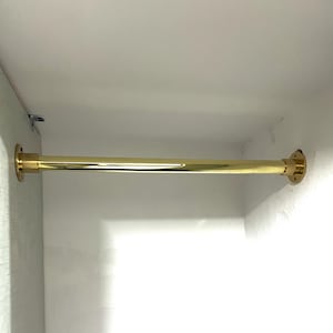 May include: A gold-coloured wardrobe rail fixed between two white surfaces. The cylindrical rail has circular mounting plates at each end. The metal rail is designed for hanging clothes, and is approximately 90 cm in length.