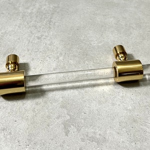 Könnte beinhalten: Ein goldener Schrankgriff mit einem durchsichtigen Acryl-Stab. Der Griff hat eine zylindrische Form und eine glatte Oberfläche.