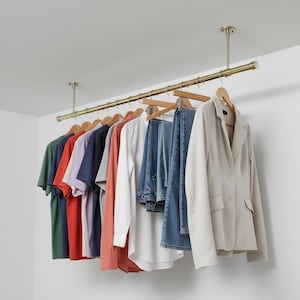 Puede incluir: Un perchero de ropa de latón suspendido del techo, que muestra una variedad de prendas. La ropa incluye camisetas en verde, rojo y morado, junto con jeans y una blazer beige. Los artículos cuelgan de perchas de madera.