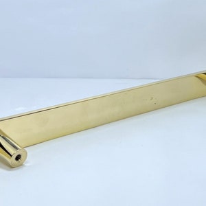 May include: A gold-colored cabinet pull with a rectangular bar and two cylindrical end pieces. The hardware is made of a shiny, metallic material and is designed for installation on a cabinet or drawer. The pull is approximately 12 inches long.