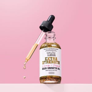 May include: A clear glass bottle with a gold label that reads "HEALTHY HAIR GROWTH SERUM TMA THE MANE ATTRACTION EXTRA STRENGTH Rosemary + Fenugreek HAIR GROWTH OIL MADE FOR ALL HAIR TYPES 2 OZ/58.1 ml". The bottle is filled with a clear liquid and has a dropper attached to the top.