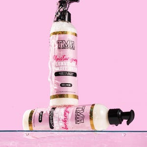 May include: Two bottles of TMA The Mane Attraction Moisturizing Leave-In Conditioner. The bottles are clear with a white liquid inside. The bottles are labeled with the brand name and product name in pink and gold lettering. The bottles are sitting in water.