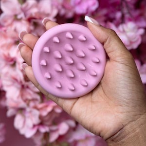 Puede incluir: Un masajeador de cuero cabelludo circular rosa claro con protuberancias redondeadas en forma de cono. El masajeador se sostiene en una mano, con un fondo borroso de flores rosas.