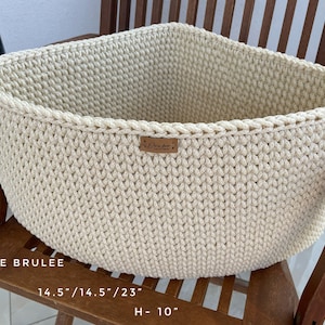 May include: A cream-colored crocheted basket with a rectangular shape. The basket is 14.5 inches wide and 10 inches tall. The basket is sitting on a wooden chair.