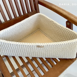 Może przedstawiać: Beżowy, trójkątny kosz do przechowywania wykonany na szydełku, umieszczony na drewnianym krześle. Kosz wykonany jest z grubego sznurka i ma plecioną fakturę.