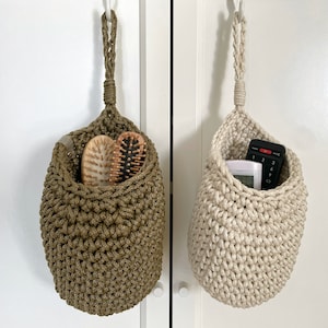 May include: Two hanging crocheted baskets, one in olive green and one in beige, with different items inside. The green basket holds two wooden hair brushes, while the beige basket holds a remote control and a small white device.
