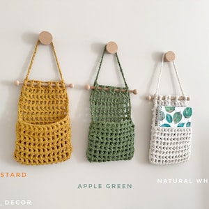 May include: Three handmade crochet wall organizers in mustard yellow, apple green, and natural white. Each has a wooden dowel and a wooden peg for hanging. The natural white organizer holds a notebook with a leaf pattern.