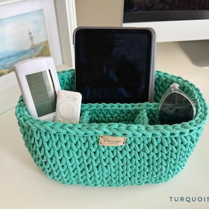 Könnte beinhalten: Ein ovaler Aufbewahrungskorb aus türkisfarbenem Häkelgarn mit internen Trennwänden und einem kleinen rechteckigen Etikett in Lederoptik mit der Aufschrift Daria decor. Der Korb enthält ein Tablet, eine weiße Fernbedienung und eine schwarze Sonnenbrille.