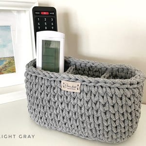 May include: A light gray crocheted basket with two compartments for storing remotes or other small items. The basket has a small label that says "Paris Love Handmade".