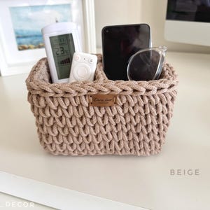 May include: Beige, hand-knitted storage basket with two compartments. The basket holds a remote control, a smartphone, and sunglasses. A small, rectangular label with text is attached to the front. The basket is on a white surface.