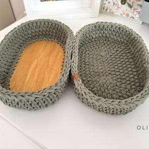 Könnte beinhalten: Zwei olivgrüne, oval geformte Häkelkörbe mit Holzsockel. Die Körbe haben eine geflochtene Textur und kleine braune Lederakzente. Das Wort "OLIVE" ist unten rechts aufgedruckt.