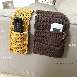 May include: Two crocheted couch armrest organizers, one in mustard yellow and one in chocolate brown. The organizers have pockets to hold remotes or other small items. The text "SMALL MUSTARD" is below the yellow organizer and the text "MEDIUM CHOCOLATE" is below the brown organizer.