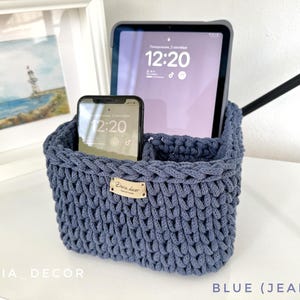 May include: A blue crochet storage basket holding a tablet and a smartphone. The tablet displays the time 12:20. The basket has a small tag that reads "Emma Luce". The text "BLUE (JEANS)" is at the bottom. The basket is made of a thick yarn.