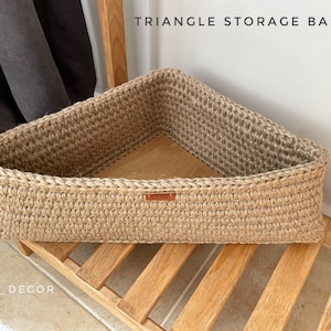 以下が含まれることがあります： 天然のジュートロープで作られた三角形の収納バスケット。バスケットはきつく均一なパターンで織られており、自然な茶色をしています。バスケットは木製の棚の上に置かれています。