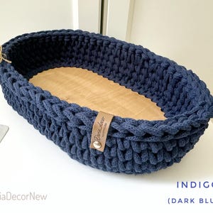 May include: A navy blue, oval-shaped crochet basket with a light wood base. The basket has a braided texture and a leather tag. The text "INDIGO (DARK BLUE)" is visible in the image. The brand name "DariaDecorNew" is also visible.