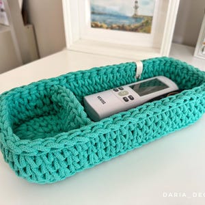 Puede incluir: Una cesta de ganchillo turquesa con forma rectangular y un divisor. Un mando a distancia blanco está dentro de la cesta. La cesta está hecha a mano y tiene un aspecto texturizado. La cesta está sobre una superficie blanca.