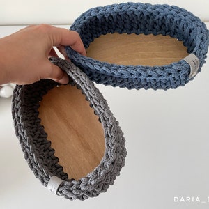 May include: Two oval-shaped crocheted baskets with wooden bottoms. The larger basket is blue and the smaller basket is gray.