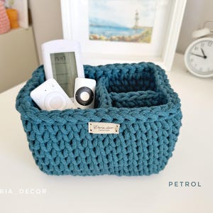 May include: A teal blue, hand-crocheted storage basket with multiple compartments, holding white remote controls. The basket has a rectangular shape and a label that reads "Daria Decor". The word "Petrol" is printed on the bottom right.