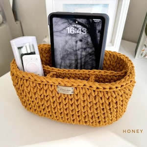 May include: A honey-colored, hand-crocheted storage basket with three compartments. The basket holds a tablet, a remote control, and a small white device. The basket has a rectangular shape with rounded edges and a small label that reads "Dora decor".