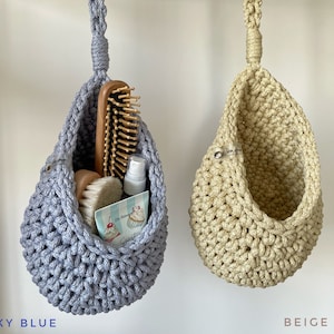May include: Two hanging crochet baskets, one sky blue and one beige, are displayed. The sky blue basket contains a wooden hairbrush, a small brush, a spray bottle, and a card. The beige basket is empty.
