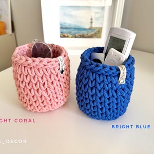 May include: Two handmade, knitted storage baskets. One is light coral, holding sunglasses, and the other is bright blue, containing remote controls. The baskets are cylindrical with a textured stitch pattern. The text "LIGHT CORAL" and "BRIGHT BLUE" are below the baskets.