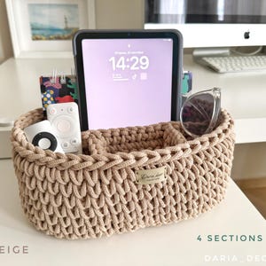 Op de afbeelding: Een beige, handgehaakte organizer mand met vier secties. De mand bevat een tablet, afstandsbedieningen, een zonnebril en een klein notitieboekje. De tekst "BEIGE" en "4 SECTIONS" zijn zichtbaar.