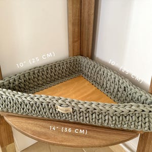 May include: A light green crocheted corner shelf with a 10 inch by 10 inch square opening and a 14 inch base. The shelf is made of thick rope and has a woven pattern.