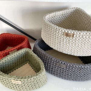 May include: Three crocheted triangular baskets in different colors: red, green, and gray. The baskets are made with a textured stitch and have a wooden base. The baskets are labeled with the text "Daria Decor".