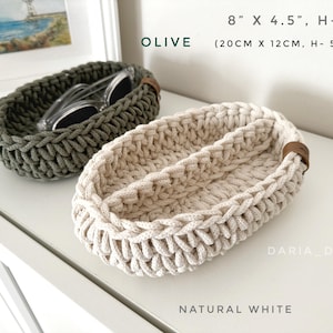 May include: Two crocheted oval-shaped baskets, one in olive green and one in natural white. The baskets are 8 inches by 4.5 inches and 2 inches tall. The baskets are made of thick cotton rope and have a leather handle.