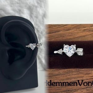 May include: A silver heart-shaped ring with a clear cubic zirconia stone and a small arrow on either side. The ring is shown on a model's ear and in a wooden box with the text "klemmmenVon" on the bottom.