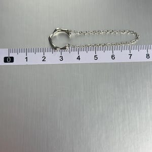 K&ouml;nnte beinhalten: Eine silberne Kettenkette mit einem einzigartigen, abstrakten Anh&auml;nger. Der Anh&auml;nger ist kreisf&ouml;rmig mit einem offenen Design. Die Kette ist etwa 18 cm lang.