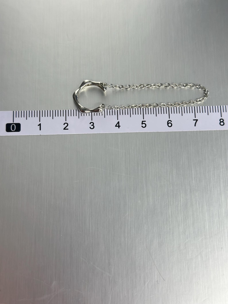 K&ouml;nnte beinhalten: Eine silberne Kettenkette mit einem einzigartigen, abstrakten Anh&auml;nger. Der Anh&auml;nger ist kreisf&ouml;rmig mit einem offenen Design. Die Kette ist etwa 18 cm lang.