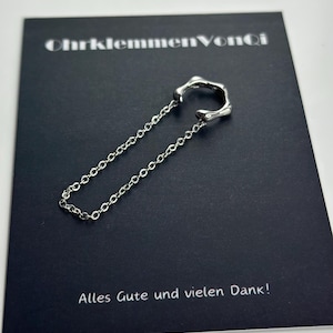 K&ouml;nnte beinhalten: Silberner Ohrclip mit einer Kette. Der Ohrclip ist kreisf&ouml;rmig mit einem gezackten Rand. Die Kette ist silberfarben und hat ein filigranes Design. Der Ohrclip liegt auf einer schwarzen Karte mit dem Text "OhrklemmenVonCi" und "Alles Gute und vielen Dank!"