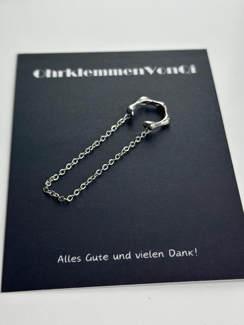K&ouml;nnte beinhalten: Silberner Ohrclip mit einer Kette. Der Ohrclip ist kreisf&ouml;rmig mit einem gezackten Rand. Die Kette ist silberfarben und hat ein filigranes Design. Der Ohrclip liegt auf einer schwarzen Karte mit dem Text "OhrklemmenVonCi" und "Alles Gute und vielen Dank!"