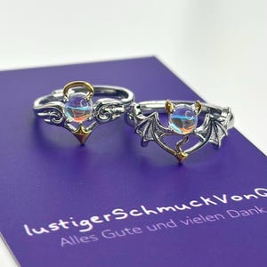 Könnte beinhalten: Zwei silberne Ringe mit schillernden Steinen. Ein Ring zeigt einen Engel mit Flügeln und einem Heiligenschein. Der andere Ring zeigt eine Fledermaus mit Flügeln und Hörnern. Die Ringe befinden sich auf einem violetten Hintergrund mit dem Text "lustiger Schmuck Von" und "Alles Gute und vielen Dank".
