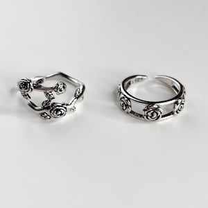 Op de afbeelding: Twee zilverkleurige ringen met rozenmotieven. De linkerring heeft een open, rankachtig ontwerp met gebeeldhouwde rozen en bladeren. De rechterring is een band met een herhalend rozenpatroon en een opening, aan de binnenzijde gestempeld met S925.